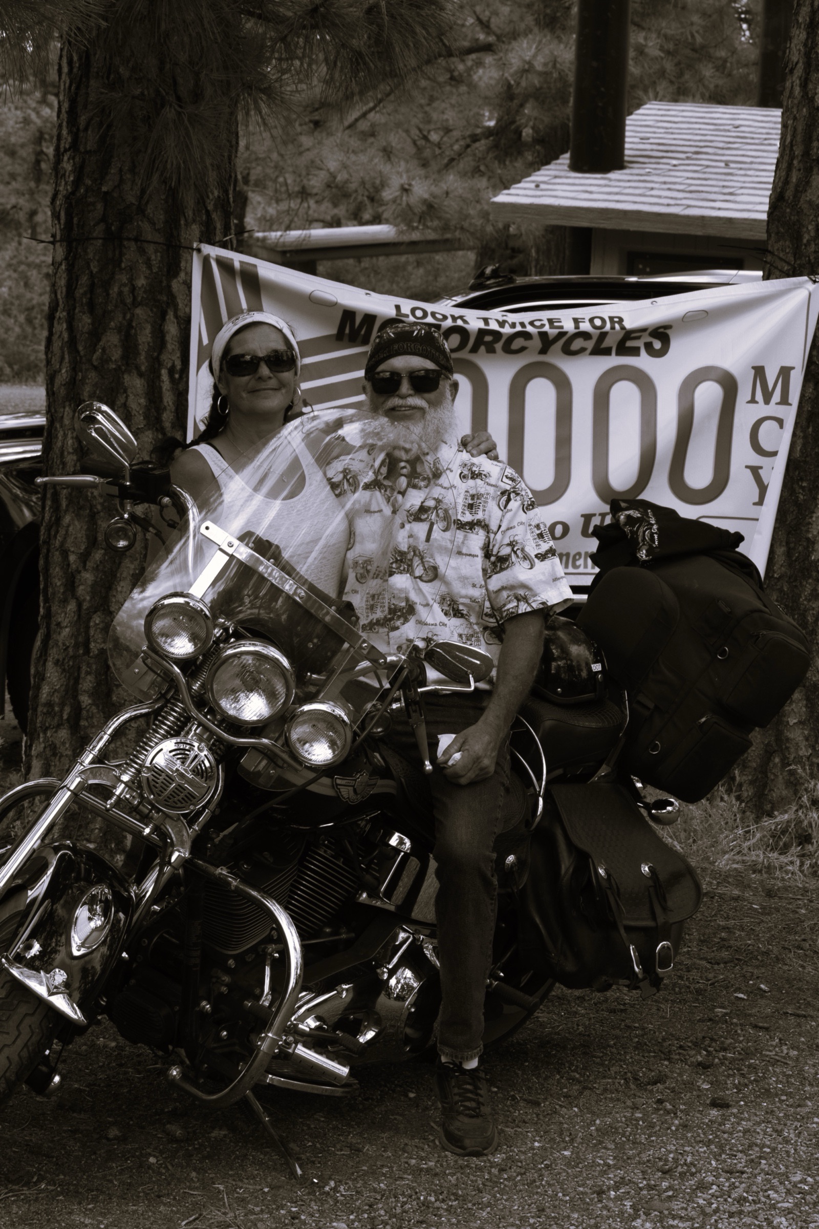 Black and white portrait of a couple sitting on a motorcycle under the trees.